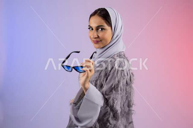 إيماءات الانوثة والجمال, الاستمتاع بامتلاك اكسسوارات عصرية, الاهتمام بالاناقة والمظهر الخارجي، سيدة اماراتية ترتدي عباءة ونظارة, بورتريه مقرب لامرأة عربية خليجية سعودية محجبة تقوم بتجربة النظارات الشمسية الجديدة, النظر إلى الكاميرا بتعابير السرور، خلفية ملونة