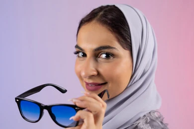 الاهتمام بالاناقة والمظهر الخارجي، سيدة اماراتية ترتدي عباءة ونظارة, الاستمتاع بامتلاك اكسسوارات عصرية, إيماءات الانوثة والجمال, بورترية مقرب لامرأة عربية خليجية سعودية محجبة تقوم بتجربة النظارات الشمسية الجديدة, النظر إلى الكاميرا بتعابير السرور، خلفية ملونة