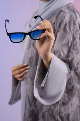 التسويق للمنتجات المحلية، عرض الأزياء والموديلات الحديثة، شراء اكسسوارات نسائية عصرية, مفهوم الانوثة والأناقة والجمال, سيدة اماراتية ترتدي عباءة رمادية, بورتريه مقرب لامرأة عربية خليجية سعودية محجبة تحمل بيدها نظارات شمسية, خلفية ملونة