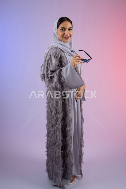 النظر إلى الكاميرا بتعابير الأنوثة والسرور, الاستمتاع بامتلاك اكسسوارات عصرية، مفهوم الأناقة والجمال, الاهتمام بالمظهر الخارجي، سيدة اماراتية مبتسمة ترتدي عباءة، بورتريه لامرأة عربية خليجية سعودية محجبة تضع نظارات شمسية, الجسم بالطول الكامل، خلفية ملونة