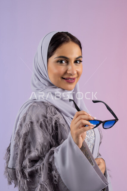 الاستمتاع بامتلاك اكسسوارات عصرية, الاهتمام بالاناقة والمظهر الخارجي، سيدة اماراتية ترتدي عباءة ونظارة, إيماءات الانوثة والجمال, بورترية مقرب لامرأة عربية خليجية سعودية محجبة تقوم بتجربة النظارات الشمسية الجديدة, النظر إلى الكاميرا بتعابير السرور، خلفية ملونة
