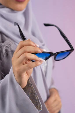 شراء اكسسوارات نسائية عصرية, التسويق للمنتجات المحلية، عرض الأزياء والموديلات الحديثة، مفهوم الانوثة والأناقة والجمال, سيدة اماراتية ترتدي عباءة رمادية, بورتريه مقرب لامرأة عربية خليجية سعودية محجبة تمسك بيدها نظارات شمسية زرقاء, خلفية ملونة