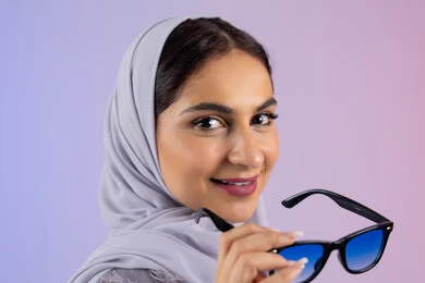 الاهتمام بالاناقة والمظهر الخارجي، سيدة اماراتية ترتدي عباءة ونظارة, الاستمتاع بامتلاك اكسسوارات عصرية, إيماءات الانوثة والجمال, بورترية مقرب لامرأة عربية خليجية سعودية محجبة تقوم بتجربة النظارات الشمسية الجديدة, النظر إلى الكاميرا بتعابير السرور، خلفية ملونة