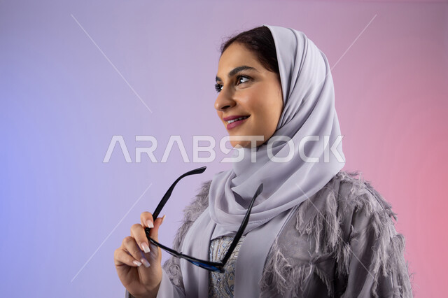 النظر إلى شيء ما بتعابير السرور، إيماءات الانوثة والدلال, الاستمتاع بامتلاك اكسسوارات عصرية, الاهتمام بالاناقة والمظهر الخارجي، سيدة اماراتية ترتدي عباءة ونظارة, بورتريه مقرب لامرأة عربية خليجية سعودية محجبة تقوم بتجربة النظارات الشمسية الجديدة, خلفية ملونة