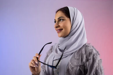 النظر إلى شيء ما بتعابير السرور، إيماءات الانوثة والدلال, الاستمتاع بامتلاك اكسسوارات عصرية, الاهتمام بالاناقة والمظهر الخارجي، سيدة اماراتية ترتدي عباءة ونظارة, بورتريه مقرب لامرأة عربية خليجية سعودية محجبة تقوم بتجربة النظارات الشمسية الجديدة, خلفية ملونة