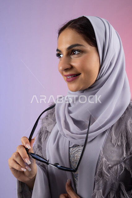 النظر إلى شيء ما بتعابير السرور، إيماءات الانوثة والدلال, الاستمتاع بامتلاك اكسسوارات عصرية, الاهتمام بالاناقة والمظهر الخارجي، سيدة اماراتية ترتدي عباءة ونظارة, بورتريه مقرب لامرأة عربية خليجية سعودية محجبة تقوم بتجربة النظارات الشمسية الجديدة, خلفية ملونة