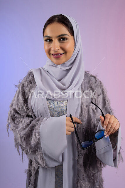 مفهوم الانوثة والأناقة والجمال, الاستمتاع بامتلاك اكسسوارات عصرية, سيدة اماراتية ترتدي عباءة ونظارة تنظر إلى الكاميرا بتعابير السرور, بورتريه لامرأة عربية خليجية سعودية محجبة تقوم بتجربة النظارات الشمسية الجديدة, خلفية ملونة