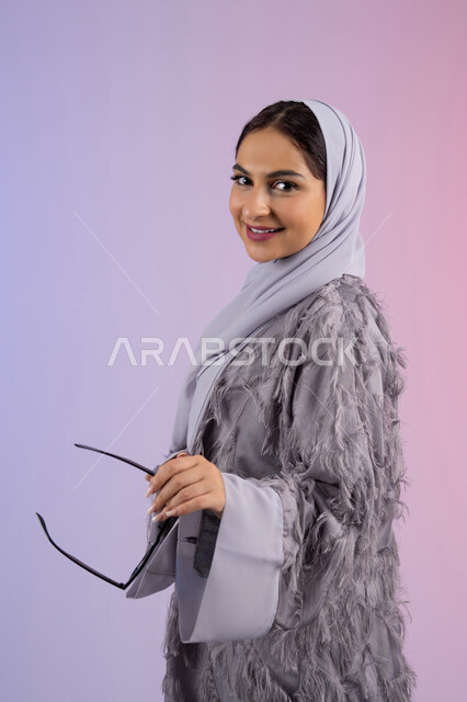 شراء اكسسوارات عصرية, مفهوم الانوثة والأناقة والجمال, سيدة اماراتية ترتدي عباءة ونظارة تنظر إلى الكاميرا بتعابير السرور, بورتريه لامرأة عربية خليجية سعودية محجبة تحمل بيدها النظارات الشمسية الجديدة, خلفية ملونة