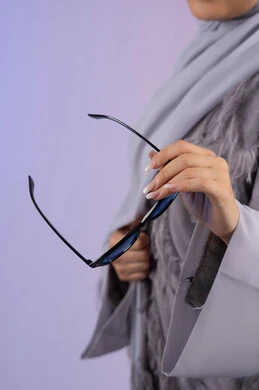شراء اكسسوارات نسائية عصرية, التسويق للمنتجات المحلية، عرض الأزياء والموديلات الحديثة، مفهوم الانوثة والأناقة والجمال, سيدة اماراتية ترتدي عباءة رمادية, بورتريه مقرب لامرأة عربية خليجية سعودية محجبة تمسك بيدها نظارات شمسية زرقاء, خلفية ملونة