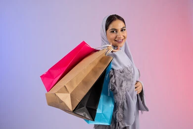 المتعة بالتسوق وشراء المستلزمات الجديدة، الأثر الايجابي للتسوق على المرأة، الترويج للمنتجات والماركات العالمية، شابة سعودية محجبة ترتدي عباءة تنظر للكاميرا بتعابير السعادة، بورتريه لامرأة عربية خليجية اماراتية تحمل بيدها أكياس تسوق ملونة، خلفية ملونة