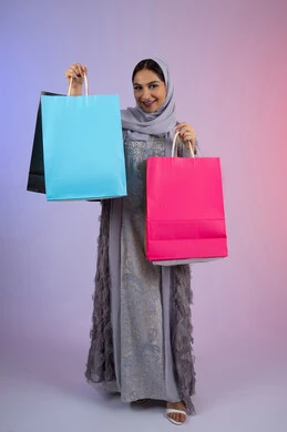 الترويج للمنتجات والماركات العالمية الفاخرة، شابة سعودية محجبة تنظر إلى الكاميرا بايماءات الإعجاب والاستمتاع بالتسوق وشراء المستلزمات الجديدة، بورتريه لامرأة عربية خليجية اماراتية ترتدي عباءة رمادية تحمل بيدها أكياس تسوق ملونة، الجسم بالطول الكامل، خلفية ملونة