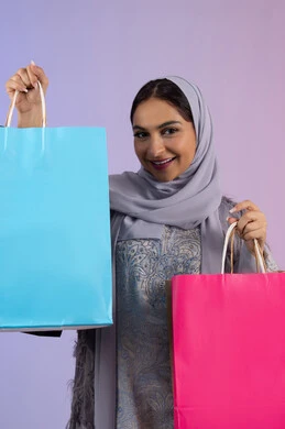 الأثر الايجابي للتسوق على المرأة، الترويج للمنتجات والماركات العالمية، شابة سعودية محجبة ترتدي عباءة تنظر للكاميرا بتعابير السعادة، الاستمتاع بالتسوق وشراء المستلزمات الجديدة، بورتريه لامرأة عربية خليجية اماراتية تحمل بيدها أكياس تسوق ملونة، خلفية ملونة