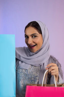 السعادة والمتعة بالتسوق وشراء المستلزمات الجديدة، الترويج للمنتجات والماركات العالمية، شابة سعودية محجبة ترتدي عباءة تنظر للكاميرا بتعابير السرور، بورتريه مقرب لامرأة عربية خليجية اماراتية تحمل بيدها أكياس تسوق ملونة، خلفية ملونة