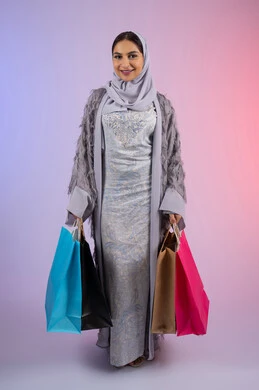 Marketing for luxury international products and brands, a young Saudi woman wearing a hijab looks at the camera with gestures of admiration and enjoyment while shopping and buying new supplies, a portrait of an Arab Gulf Emirati woman wearing a gray abaya and holding colorful shopping bags, full body, colorful background