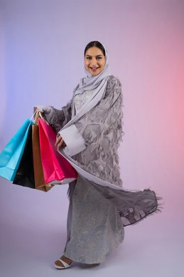 الترويج للمنتجات والماركات العالمية الفاخرة، شابة سعودية محجبة تنظر إلى الكاميرا بايماءات الإعجاب والاستمتاع بالتسوق وشراء المستلزمات الجديدة، بورتريه لامرأة عربية خليجية اماراتية ترتدي عباءة رمادية تحمل بيدها أكياس تسوق ملونة، الجسم بالطول الكامل، خلفية ملونة
