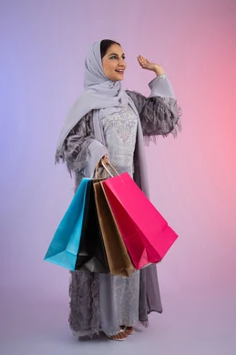 رفع اليد بإيماءات القاء التحية والسلام، التسويق للمنتجات والماركات العالمية الفاخرة، شابة سعودية محجبة ترتدي عباءة، الاستمتاع بالتسوق وشراء المستلزمات الجديدة، بورتريه لامرأة عربية خليجية اماراتية تحمل بيدها أكياس تسوق ملونة، الجسم بالطول الكامل، خلفية ملونة