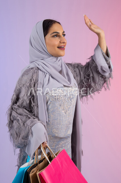 الأثر الايجابي للتسوق على المرأة، رفع اليد بإيماءات إلقاء السلام، التسويق للمنتجات والماركات العالمية، شابة سعودية محجبة ترتدي عباءة، الاستمتاع بالتسوق وشراء المستلزمات الجديدة، بورتريه لامرأة عربية خليجية اماراتية تحمل بيدها أكياس تسوق ملونة، خلفية ملونة