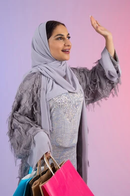 الأثر الايجابي للتسوق على المرأة، رفع اليد بإيماءات إلقاء السلام، التسويق للمنتجات والماركات العالمية، شابة سعودية محجبة ترتدي عباءة، الاستمتاع بالتسوق وشراء المستلزمات الجديدة، بورتريه لامرأة عربية خليجية اماراتية تحمل بيدها أكياس تسوق ملونة، خلفية ملونة