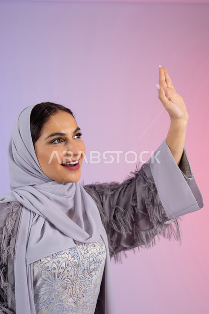 تعابير السعادة بمقابلة شخص ما، الاناقة والاهتمام بالمظهر الخارجي، شابة سعودية محجبة ترتدي عباءة تنظر إلى شيء ما بإيماءات السعادة والسرور، بورتريه مقرب لامرأة عربية خليجية اماراتية ترفع يدها بإيماءات إلقاء التحية والسلام، خلفية ملونة