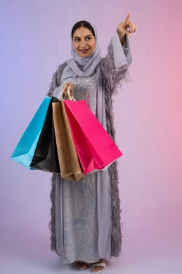 الترويج للمنتجات والماركات العالمية، الإشارة بإصبع السبابة إلى شيء ما، الأثر الايجابي للتسوق على المرأة، شابة سعودية محجبة ترتدي عباءة، الاستمتاع بالتسوق وشراء المستلزمات الجديدة، بورتريه لامرأة عربية خليجية اماراتية تحمل بيدها أكياس تسوق ملونة، خلفية ملونة