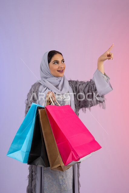 الإشارة بإصبع السبابة إلى شيء ما، الأثر الايجابي للتسوق على المرأة، التسويق للمنتجات والماركات العالمية، شابة سعودية محجبة ترتدي عباءة، الاستمتاع بالتسوق وشراء المستلزمات الجديدة، بورتريه لامرأة عربية خليجية اماراتية تحمل بيدها أكياس تسوق ملونة، خلفية ملونة