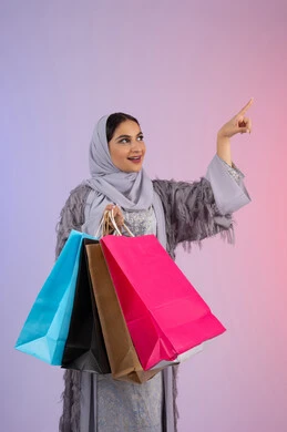 الإشارة بإصبع السبابة إلى شيء ما، الأثر الايجابي للتسوق على المرأة، التسويق للمنتجات والماركات العالمية، شابة سعودية محجبة ترتدي عباءة، الاستمتاع بالتسوق وشراء المستلزمات الجديدة، بورتريه لامرأة عربية خليجية اماراتية تحمل بيدها أكياس تسوق ملونة، خلفية ملونة