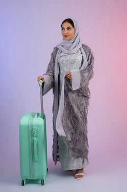 الاستعداد لقضاء رحلة صيفية ممتعة, السياحة في إجازة الصيف, بورتريه لشابة عربية خليجية سعودية مبتسمة محجبة ترتدي العباءة تمسك حقيبة السفر ، امرأة اماراتية تقف بإيماءات الثقة بالنفس, الشعور بالسعادة والفرح، الجسم بالطول الكامل, خلفية ملونة.