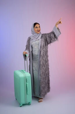 الاستعداد لقضاء رحلة صيفية ممتعة, الاشارة بإصبع السبابة إلى شيء ما، تجهيز حقائب الامتعة، السياحة في إجازة الصيف, بورتريه لشابة عربية خليجية سعودية مبتسمة ترتدي العباءة تمسك حقيبة السفر، امرأة اماراتية محجبة تقف بإيماءات السرور, طول الجسم بالكامل، خلفية ملونة.