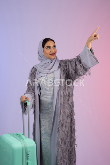 الاشارة بإصبع السبابة إلى شيء ما، تجهيز حقائب الامتعة، السياحة في إجازة الصيف, الاستعداد لقضاء رحلة صيفية ممتعة, بورتريه لشابة عربية خليجية سعودية مبتسمة ترتدي العباءة تمسك حقيبة السفر، امرأة اماراتية محجبة تقف بإيماءات السرور, خلفية ملونة.