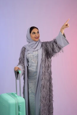 الاشارة بإصبع السبابة إلى شيء ما، تجهيز حقائب الامتعة، السياحة في إجازة الصيف, الاستعداد لقضاء رحلة صيفية ممتعة, بورتريه لشابة عربية خليجية سعودية مبتسمة ترتدي العباءة تمسك حقيبة السفر، امرأة اماراتية محجبة تقف بإيماءات السرور, خلفية ملونة.