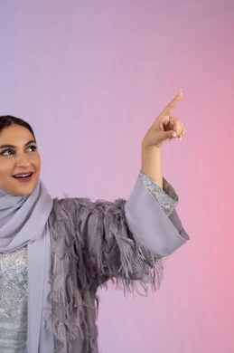 تعابير السعادة برؤية شيء ما، الاناقة والاهتمام بالمظهر الخارجي، شابة سعودية محجبة ترتدي عباءة تنظر إلى شيء ما بإيماءات السعادة والسرور، بورتريه مقرب لامرأة عربية خليجية اماراتية تشير بإصبع السبابة إلى شيء ما، خلفية ملونة