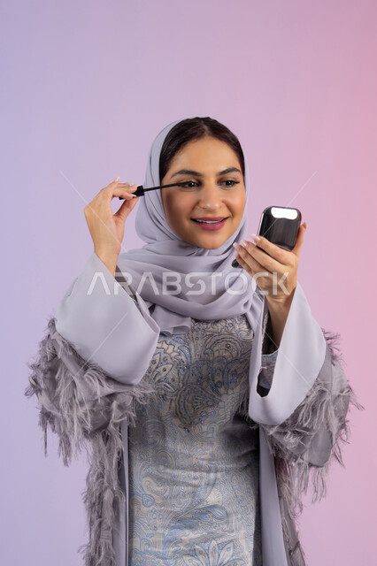 The concept of adornment and beautification, interest in beauty and outward appearance, passion for applying makeup, promoting cosmetics and products, close-up portrait of a veiled Emirati Gulf Arab woman wearing an abaya applying mascara, a young Saudi woman applying makeup, colorful background
