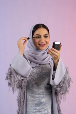 مفهوم التزين والتجمل, الاهتمام بالجمال والمظهر الخارجي, الشغف بوضع الميك اب، الترويج لمنتجات ومستحضرات التجميل, بورتريه مقرب لامرأة عربية خليجية إماراتية محجبة ترتدي العباءة تقوم باستخدام المسكرة, شابة سعودية تضع المكياج, خلفية ملونة