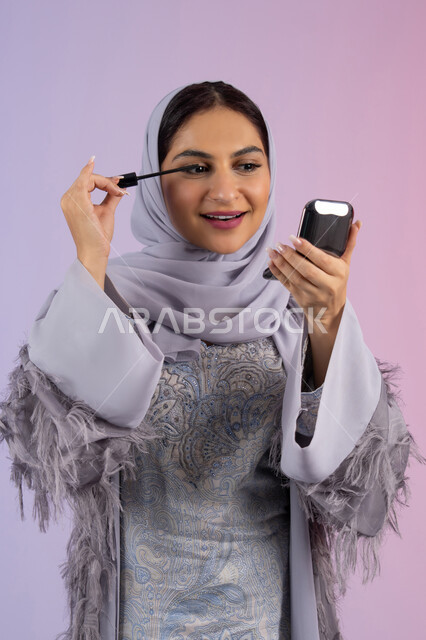 الشغف بوضع الميك اب، مفهوم التزين والتجمل, الاهتمام بالجمال والمظهر الخارجي, الترويج لمنتجات ومستحضرات التجميل, بورتريه مقرب لامرأة عربية خليجية إماراتية محجبة ترتدي العباءة تقوم باستخدام المسكرة, شابة سعودية تضع المكياج, خلفية ملونة