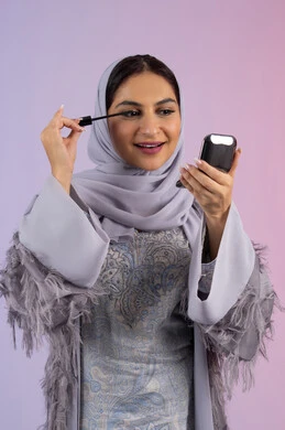 الشغف بوضع الميك اب، مفهوم التزين والتجمل, الاهتمام بالجمال والمظهر الخارجي, الترويج لمنتجات ومستحضرات التجميل, بورتريه مقرب لامرأة عربية خليجية إماراتية محجبة ترتدي العباءة تقوم باستخدام المسكرة, شابة سعودية تضع المكياج, خلفية ملونة