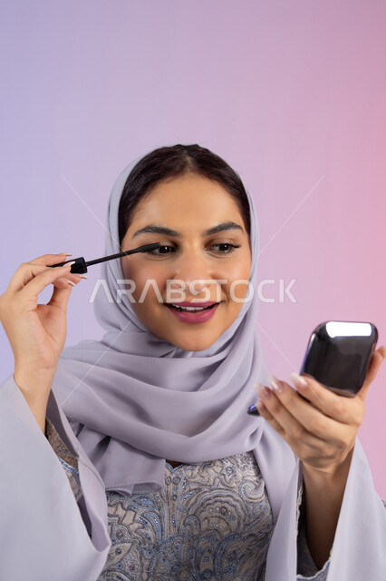 الترويج لمنتجات ومستحضرات التجميل, الشغف بوضع الميك اب، مفهوم التزين والتجمل, الاهتمام بالجمال والمظهر الخارجي, بورتريه مقرب لامرأة عربية خليجية إماراتية محجبة ترتدي العباءة تقوم باستخدام المسكرة, شابة سعودية تضع المكياج, خلفية ملونة