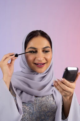 الترويج لمنتجات ومستحضرات التجميل, الشغف بوضع الميك اب، مفهوم التزين والتجمل, الاهتمام بالجمال والمظهر الخارجي, بورتريه مقرب لامرأة عربية خليجية إماراتية محجبة ترتدي العباءة تقوم باستخدام المسكرة, شابة سعودية تضع المكياج, خلفية ملونة