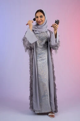 الاستمتاع بوضع الميك اب، الاهتمام بالجمال والمظهر الخارجي, التسويق لمساحيق التجميل, مفهوم التزين والتجمل, بورتريه لامرأة عربية خليجية إماراتية محجبة ترتدي العباءة تقوم باستخدام فرشاة أحمر الخدود, شابة سعودية تضع مكياج تتوريال, الجسم بالطول الكامل، خلفية ملونة