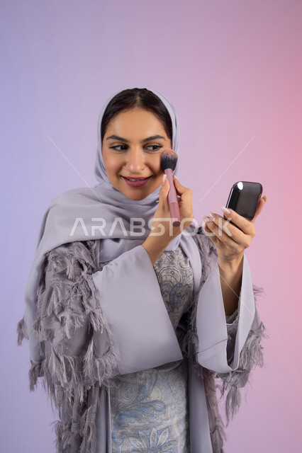 التسويق لمنتجات ومستحضرات التجميل المحلية والعالمية, الشغف بوضع الميك اب، مفهوم التزين والتجمل, الاهتمام بالجمال والمظهر الخارجي, بورتريه لامرأة عربية خليجية إماراتية محجبة ترتدي العباءة تقوم باستخدام فرشاة أحمر الخدود, شابة سعودية تضع مكياج تتوريال, خلفية ملونة
