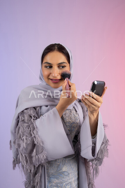 التسويق لمنتجات ومستحضرات التجميل المحلية والعالمية, الشغف بوضع الميك اب، مفهوم التزين والتجمل, الاهتمام بالجمال والمظهر الخارجي, بورتريه لامرأة عربية خليجية إماراتية محجبة ترتدي العباءة تقوم باستخدام فرشاة أحمر الخدود, شابة سعودية تضع مكياج تتوريال, خلفية ملونة