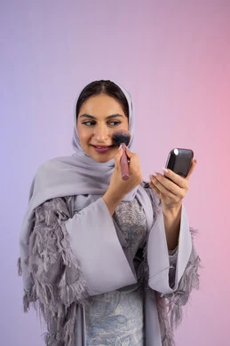 التسويق لمنتجات ومستحضرات التجميل المحلية والعالمية, الشغف بوضع الميك اب، مفهوم التزين والتجمل, الاهتمام بالجمال والمظهر الخارجي, بورتريه لامرأة عربية خليجية إماراتية محجبة ترتدي العباءة تقوم باستخدام فرشاة أحمر الخدود, شابة سعودية تضع مكياج تتوريال, خلفية ملونة