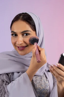 الاهتمام بالجمال والمظهر الخارجي, التسويق لمنتجات ومستحضرات التجميل المحلية, الشغف بوضع الميك اب، مفهوم التزين والتجمل, بورتريه مقرب لامرأة عربية خليجية إماراتية محجبة ترتدي العباءة تقوم باستخدام فرشاة أحمر الخدود, شابة سعودية تضع مكياج تتوريال, خلفية ملونة