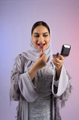 الاناقة والتزين والتجمل, احتراف واتقان وضع الميك اب، الاهتمام بالجمال والمظهر الخارجي, التسويق لمساحيق التجميل, بورتريه مقرب لامرأة عربية خليجية إماراتية محجبة ترتدي العباءة تقوم باستخدام قلم أحمر الشفاه, شابة سعودية تضع مكياج تتوريال, خلفية ملونة