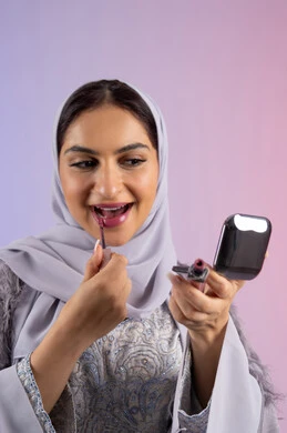 الاناقة والتزين والتجمل, احتراف واتقان وضع الميك اب، الاهتمام بالجمال والمظهر الخارجي, التسويق لمساحيق التجميل, بورتريه مقرب لامرأة عربية خليجية إماراتية محجبة ترتدي العباءة تقوم باستخدام قلم أحمر الشفاه, شابة سعودية تضع مكياج تتوريال, خلفية ملونة