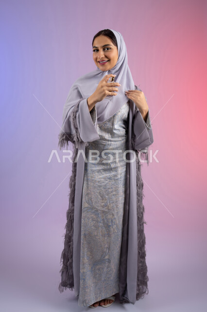 عطور عربية خليجية، الترويج للعطور والزيوت الطبيعية المحلية، مفهوم التعطر والتطيب، استخدام العود العطري، سيدة اماراتية تسوق للمنتجات العطرية، بورتريه لامرأه عربيه سعودية محجبة ترتدي العباءة تقوم برش العطر، الجسم بالطول الكامل، خلفية ملونة