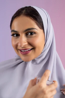 التسويق للعطور والزيوت الطبيعية العالمية، مفهوم التعطر والتطيب، استخدام العود العطري، عطورات عربية خليجية، سيدة اماراتية تسوق للمنتجات العطرية، بورتريه مقرب لامرأه عربيه سعودية محجبة ترتدي العباءة تقوم برش العطر، خلفية ملونة