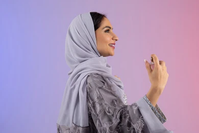 التسويق للعطور والزيوت الطبيعية العالمية، مفهوم التعطر والتطيب، استخدام العود العطري، عطورات عربية خليجية، سيدة اماراتية تسوق للمنتجات العطرية، بورتريه مقرب لامرأه عربيه سعودية محجبة ترتدي العباءة تقوم برش العطر، خلفية ملونة
