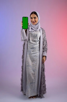 النظر إلى الكاميرا بتعابير السرور والاعجاب، عرض تطبيقات الجوال عبر شاشة خضراء فارغة كروما، بورتريه لامرأة عربية خليجية اماراتية محجبة ترتدي العباءة تمسك بيدها الهاتف المحمول، شابة سعودية تستخدم جهاز تقني حديث ومتطور، الجسم بالطول الكامل، خلفية ملونة