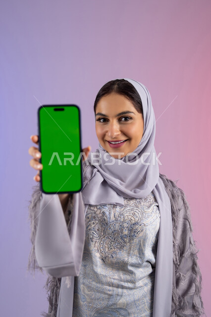 عرض تطبيقات الجوال عبر شاشة خضراء فارغة كروما، بورتريه مقرب لامرأة عربية خليجية اماراتية محجبة ترتدي العباءة تمسك بيدها الهاتف المحمول، شابة سعودية تستخدم جهاز تقني حديث ومتطور، النظر إلى الكاميرا بتعابير السرور والاعجاب، خلفية ملونة