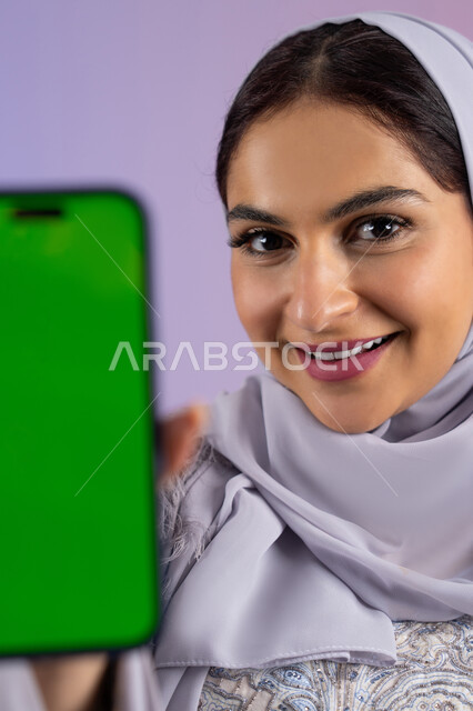 Mobile application display on blank green screen chroma key, close-up portrait of veiled Emirati Gulf Arab woman wearing abaya holding mobile phone, young Saudi woman using modern and advanced technical device, looking at camera with expressions of pleasure and admiration, colorful background